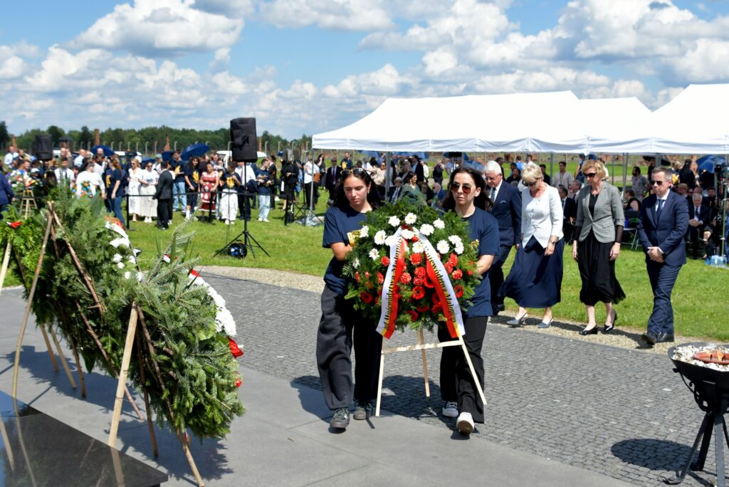 Uczestnicy uroczystości upamiętniającej zagładę Romów i Sinti składają wieniec przy pomniku w byłym obozie Auschwitz II-Birkenau. W tle widoczne namioty, zgromadzeni goście i przedstawiciele władz.