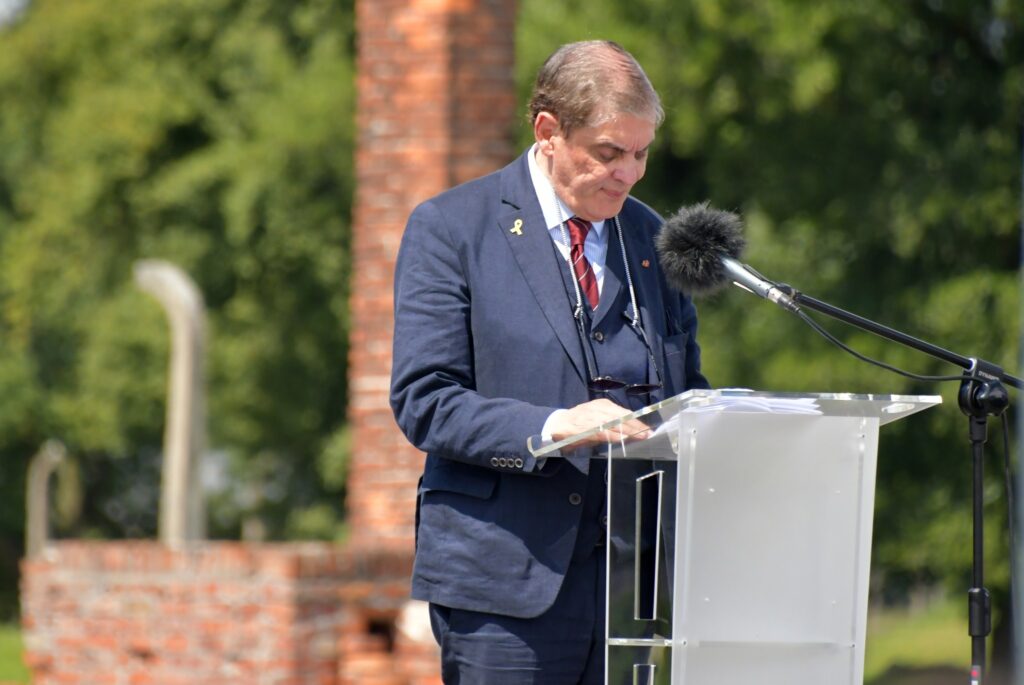 Uczestnicy uroczystości upamiętniającej zagładę Romów i Sinti składają wieniec przy pomniku w byłym obozie Auschwitz II-Birkenau. W tle widoczne namioty, zgromadzeni goście i przedstawiciele władz.