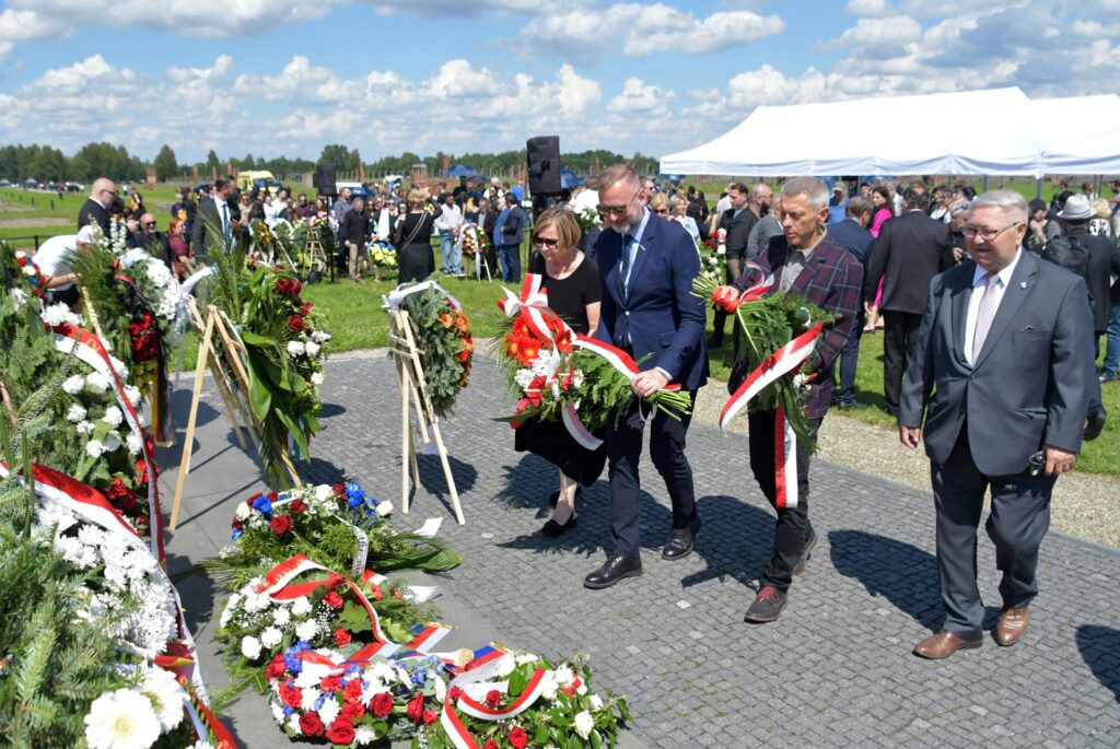 Uczestnicy uroczystości upamiętniającej zagładę Romów i Sinti składają wieniec przy pomniku w byłym obozie Auschwitz II-Birkenau. W tle widoczne namioty, zgromadzeni goście i przedstawiciele władz.