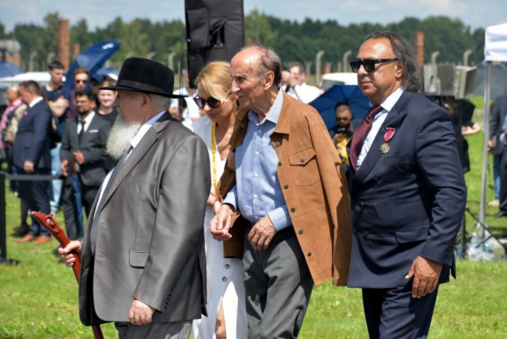 Uczestnicy uroczystości upamiętniającej zagładę Romów i Sinti składają wieniec przy pomniku w byłym obozie Auschwitz II-Birkenau. W tle widoczne namioty, zgromadzeni goście i przedstawiciele władz.