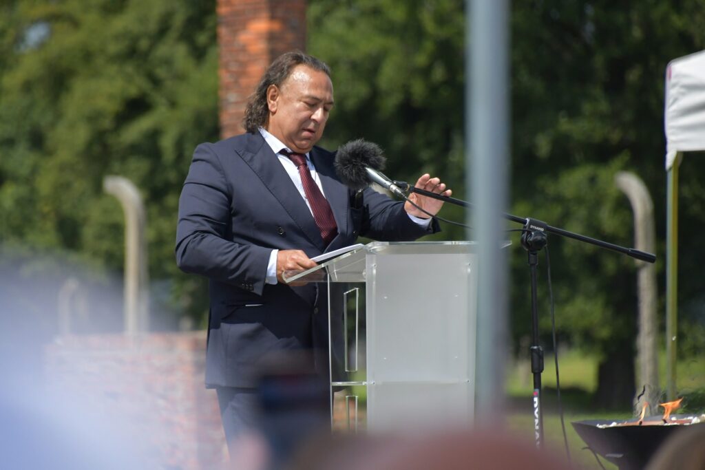Uczestnicy uroczystości upamiętniającej zagładę Romów i Sinti składają wieniec przy pomniku w byłym obozie Auschwitz II-Birkenau. W tle widoczne namioty, zgromadzeni goście i przedstawiciele władz.