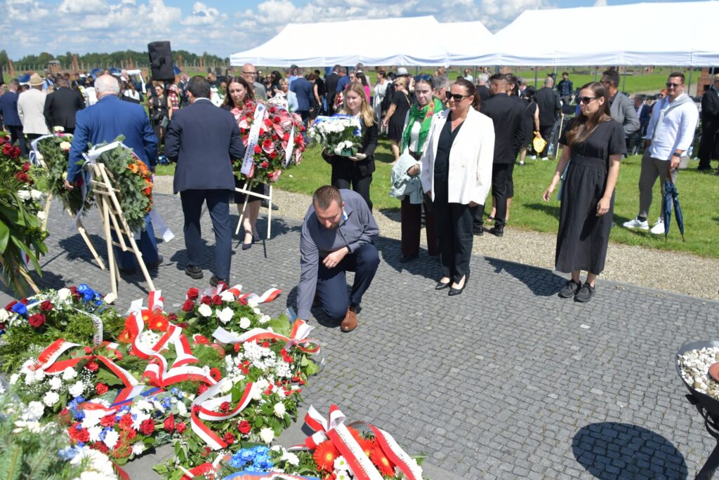 Uczestnicy uroczystości upamiętniającej zagładę Romów i Sinti składają wieniec przy pomniku w byłym obozie Auschwitz II-Birkenau. W tle widoczne namioty, zgromadzeni goście i przedstawiciele władz.
