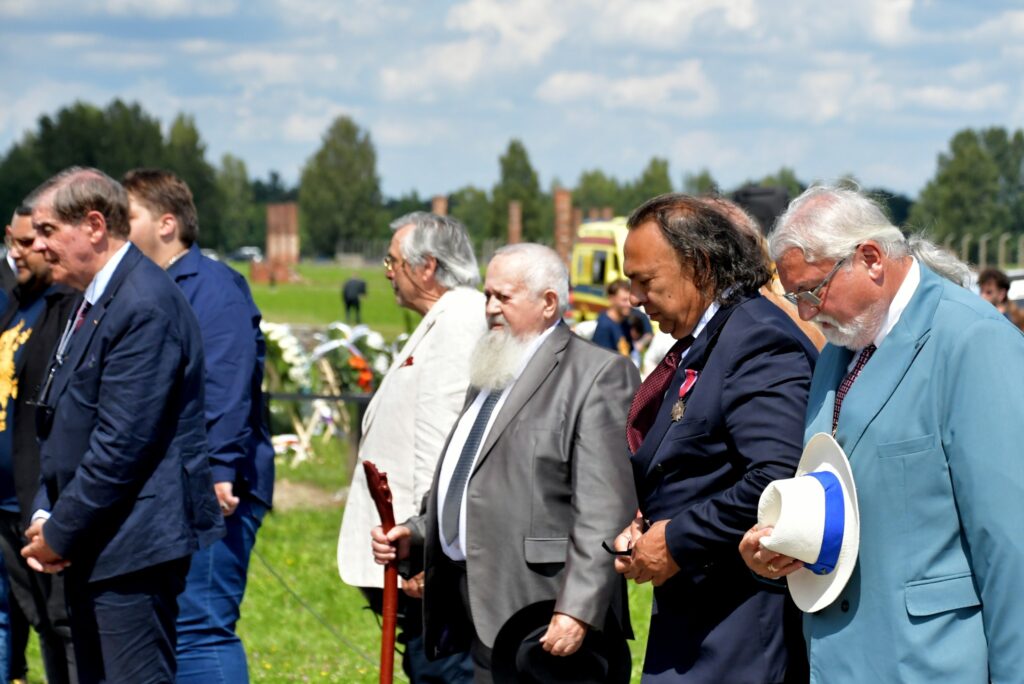 Uczestnicy uroczystości upamiętniającej zagładę Romów i Sinti składają wieniec przy pomniku w byłym obozie Auschwitz II-Birkenau. W tle widoczne namioty, zgromadzeni goście i przedstawiciele władz.