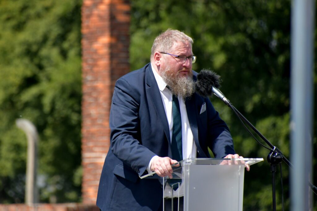 Uczestnicy uroczystości upamiętniającej zagładę Romów i Sinti składają wieniec przy pomniku w byłym obozie Auschwitz II-Birkenau. W tle widoczne namioty, zgromadzeni goście i przedstawiciele władz.