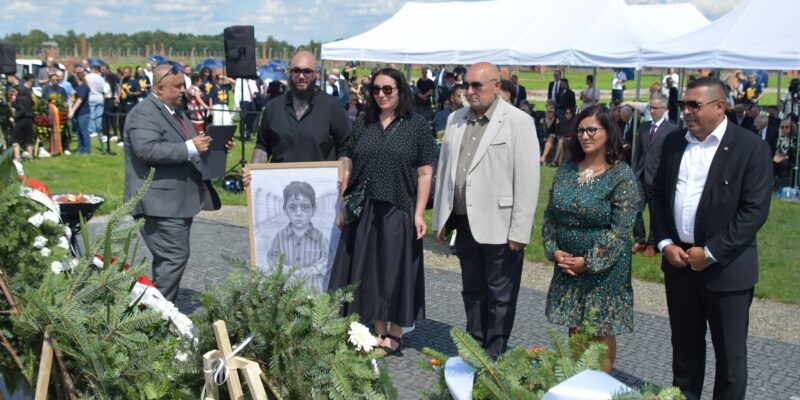 Uczestnicy uroczystości upamiętniającej zagładę Romów i Sinti składają wieniec przy pomniku w byłym obozie Auschwitz II-Birkenau. W tle widoczne namioty, zgromadzeni goście i przedstawiciele władz.