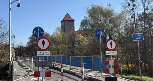 Kładka pieszo-rowerowa przy zamku w Oświęcimiu z ograniczeniami ruchu – znaki zakazu, tymczasowe barierki i zwężone przejście, w tle wieża zamkowa i rzeka Soła.