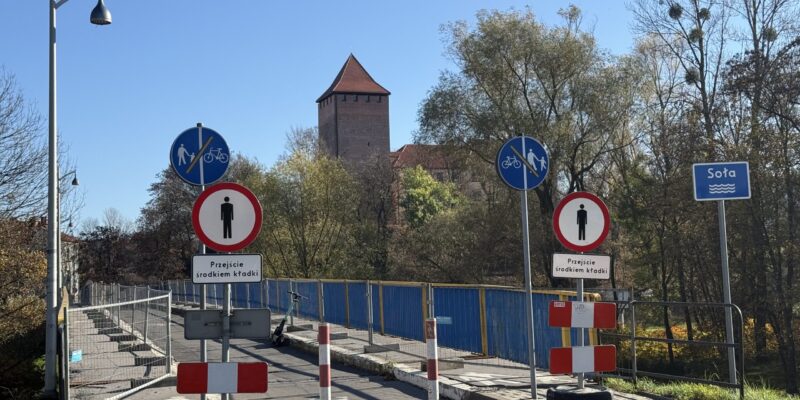 Kładka pieszo-rowerowa przy zamku w Oświęcimiu z ograniczeniami ruchu – znaki zakazu, tymczasowe barierki i zwężone przejście, w tle wieża zamkowa i rzeka Soła.