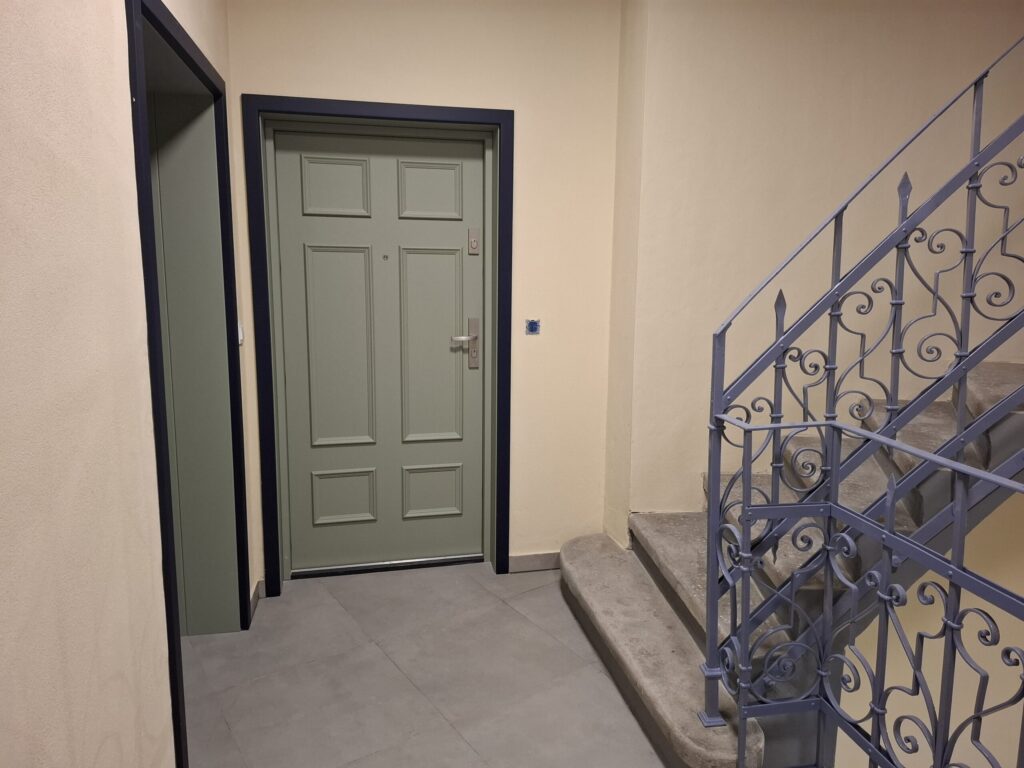 Odnowiona zabytkowa kamienica przy ul. Mickiewicza w Oświęcimiu, z elegancką jasną elewacją, zielonymi balkonami i drewnianymi oknami, w otoczeniu drzew i błękitnego nieba.