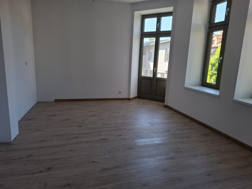 Odnowiona zabytkowa kamienica przy ul. Mickiewicza w Oświęcimiu, z elegancką jasną elewacją, zielonymi balkonami i drewnianymi oknami, w otoczeniu drzew i błękitnego nieba.