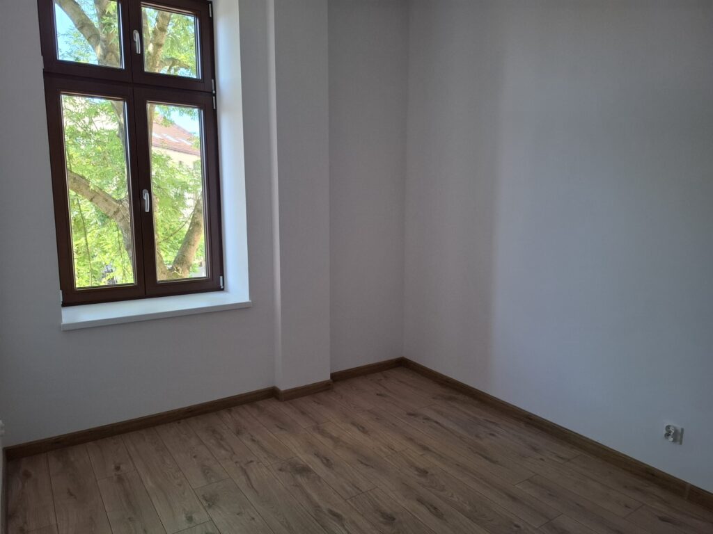 Odnowiona zabytkowa kamienica przy ul. Mickiewicza w Oświęcimiu, z elegancką jasną elewacją, zielonymi balkonami i drewnianymi oknami, w otoczeniu drzew i błękitnego nieba.