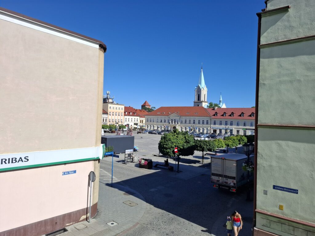 Odnowiona zabytkowa kamienica przy ul. Mickiewicza w Oświęcimiu, z elegancką jasną elewacją, zielonymi balkonami i drewnianymi oknami, w otoczeniu drzew i błękitnego nieba.