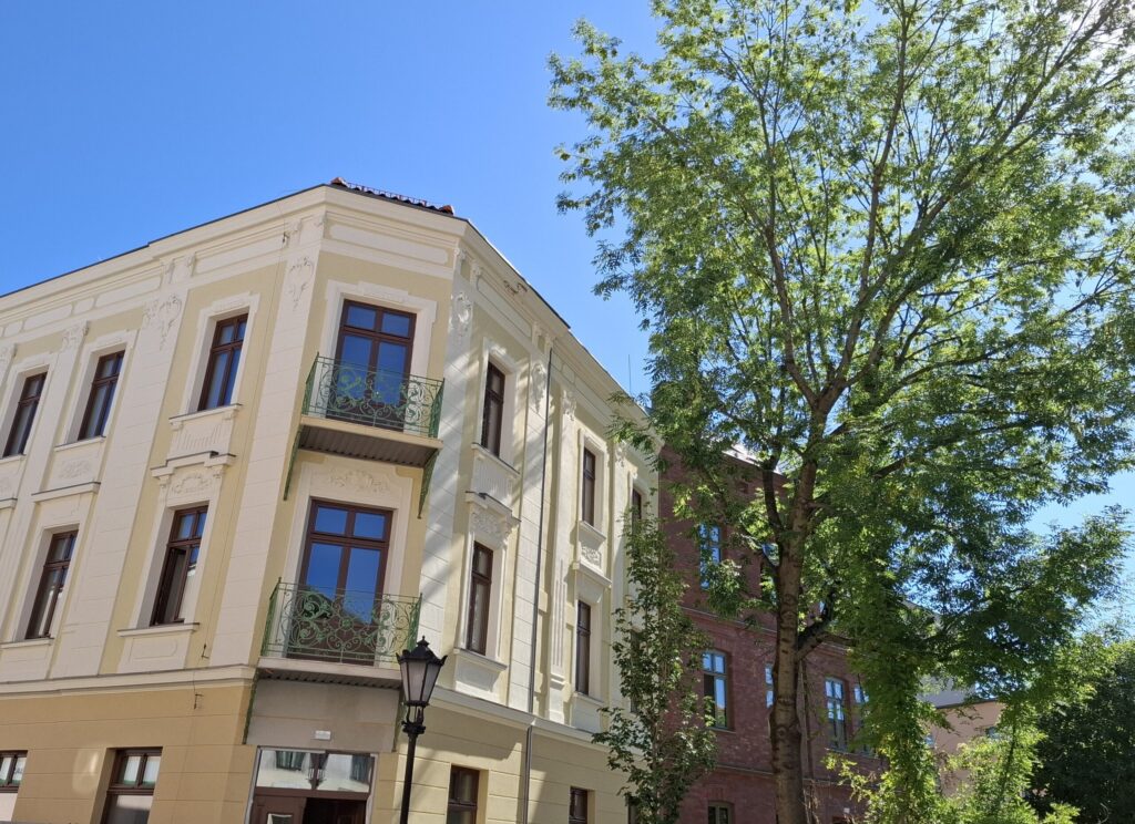 Odnowiona zabytkowa kamienica przy ul. Mickiewicza w Oświęcimiu, z elegancką jasną elewacją, zielonymi balkonami i drewnianymi oknami, w otoczeniu drzew i błękitnego nieba.
