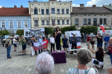 Grupa osób zgromadzona na rynku w Oświęcimiu podczas protestu społecznego z transparentem „Stop imigrantom”. Kobieta przemawia ze sceny ustawionej na podeście, wokół widoczni uczestnicy wydarzenia, flagi Polski i banery organizatorów. W tle historyczna zabudowa rynku.