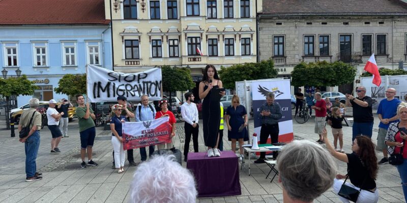 Grupa osób zgromadzona na rynku w Oświęcimiu podczas protestu społecznego z transparentem „Stop imigrantom”. Kobieta przemawia ze sceny ustawionej na podeście, wokół widoczni uczestnicy wydarzenia, flagi Polski i banery organizatorów. W tle historyczna zabudowa rynku.
