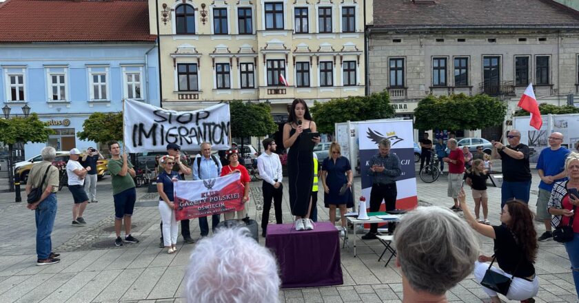 Grupa osób zgromadzona na rynku w Oświęcimiu podczas protestu społecznego z transparentem „Stop imigrantom”. Kobieta przemawia ze sceny ustawionej na podeście, wokół widoczni uczestnicy wydarzenia, flagi Polski i banery organizatorów. W tle historyczna zabudowa rynku.
