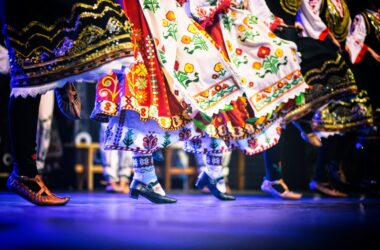 Tancerze w tradycyjnych strojach ludowych wykonują folklorystyczny taniec sceniczny; widoczne barwne haftowane spódnice, skarpety i buty charakterystyczne dla różnych regionów Europy.