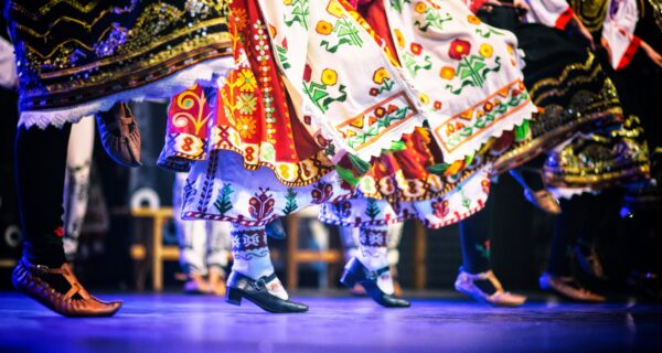 Tancerze w tradycyjnych strojach ludowych wykonują folklorystyczny taniec sceniczny; widoczne barwne haftowane spódnice, skarpety i buty charakterystyczne dla różnych regionów Europy.