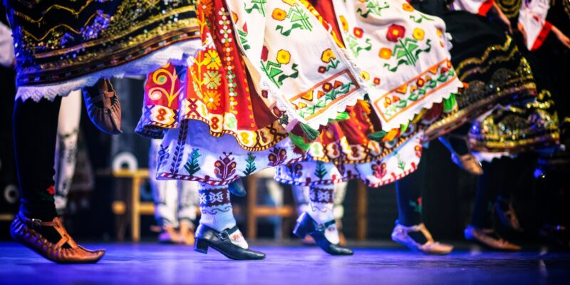 Tancerze w tradycyjnych strojach ludowych wykonują folklorystyczny taniec sceniczny; widoczne barwne haftowane spódnice, skarpety i buty charakterystyczne dla różnych regionów Europy.