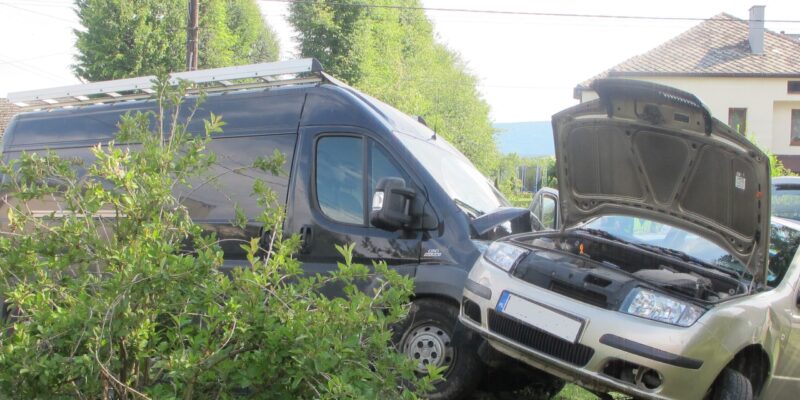 Kolizja drogowa – srebrny samochód osobowy z otwartą maską oparty o bok czarnego samochodu dostawczego, oba pojazdy znajdują się na trawniku przy domach jednorodzinnych.