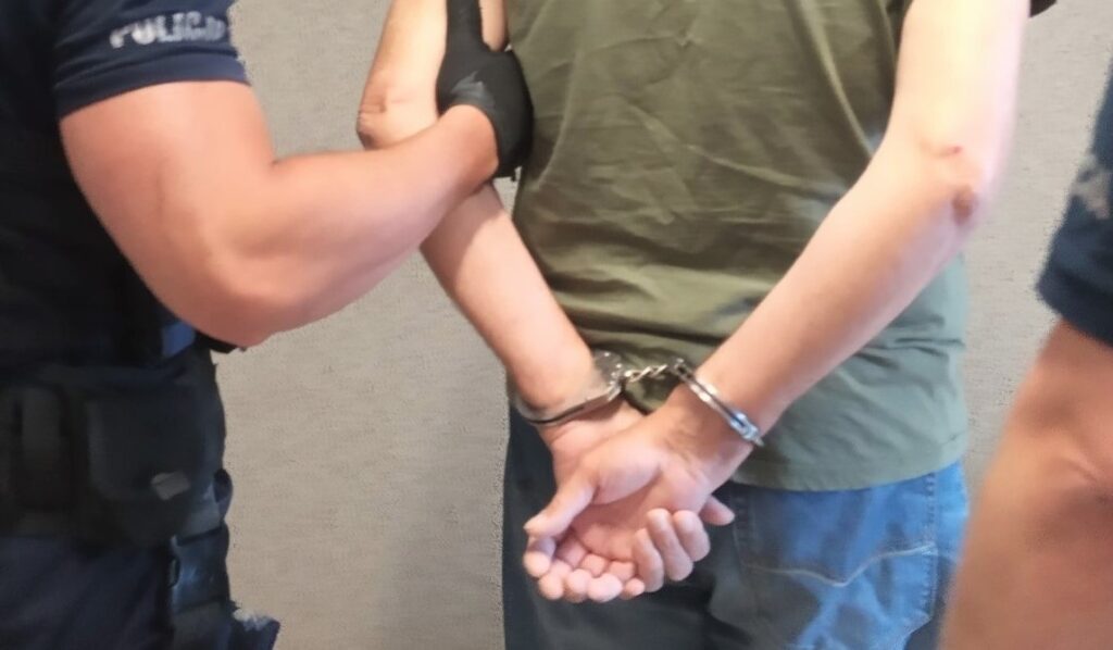 Policjant zatrzymuje mężczyznę w zielonej koszulce i kajdankach założonych na ręce trzymane z tyłu – widok z tyłu podczas interwencji.