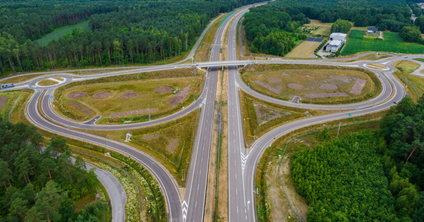 Węzeł drogowy Wola na budowanej trasie ekspresowej S1 między Kosztowami a Oświęcimiem. Zdjęcie lotnicze przedstawia nową infrastrukturę drogową otoczoną lasem i terenami wiejskimi. Widoczne są jezdnie, ronda, estakady oraz pasy włączeń i zjazdów. Data wykonania zdjęcia: 8 lipca 2025 roku.
