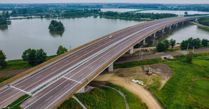 Nowo wybudowany węzeł drogowy na drodze ekspresowej S-1 w pobliżu Oświęcimia, widziany z lotu ptaka. Zdjęcie przedstawia skrzyżowanie wielopoziomowe z nowoczesnymi jezdniami, barierami energochłonnymi, ekranami akustycznymi i oznakowaniem kierunkowym. Otoczenie stanowią pola uprawne i lasy. Data wykonania zdjęcia: 25 lipca 2025 roku.
