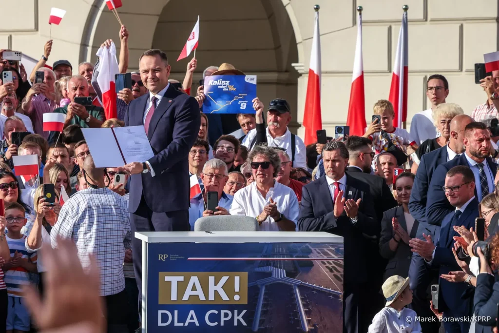 Prezydent Karol Nawrocki stoi na podium podczas spotkania w Kaliszu, trzyma w rękach podpisany projekt ustawy o realizacji inwestycji Centralnego Portu Komunikacyjnego. Za nim tłum ludzi z biało-czerwonymi flagami, część robi zdjęcia telefonami. Na pulpicie przed prezydentem napis: „TAK! DLA CPK” oraz zdjęcie lotniska. W tle widoczne polskie flagi na masztach