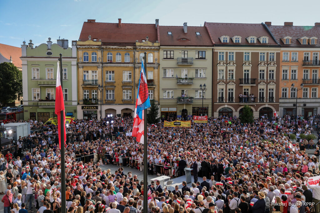 Widok z góry na tłum zgromadzony na rynku w Kaliszu podczas przemówienia prezydenta Karola Nawrockiego. W centrum ustawione podium, wokół setki osób z biało-czerwonymi flagami. W tle kamienice z balkonami, na których również stoją obserwatorzy.
