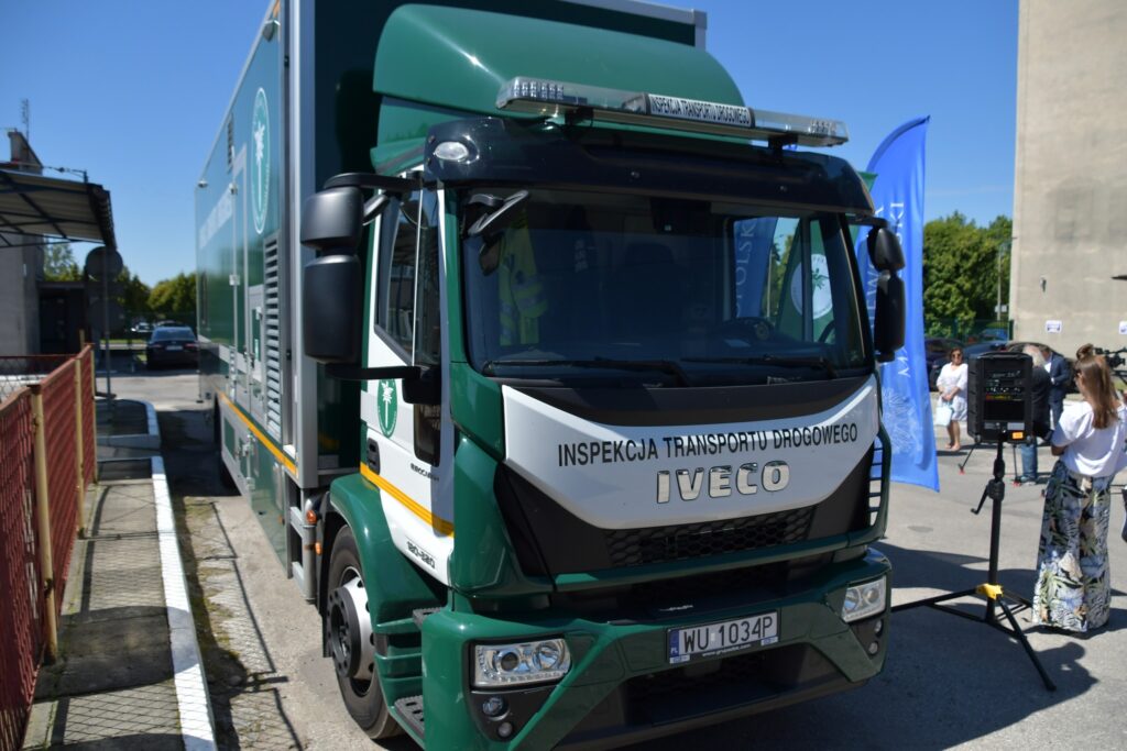 Ciężarowy pojazd Inspekcji Transportu Drogowego marki Iveco zaparkowany na placu, obok którego ustawiono sprzęt nagłośnieniowy i banery promocyjne. W tle widoczni uczestnicy wydarzenia plenerowego oraz fragment budynku i ogrodzenia.