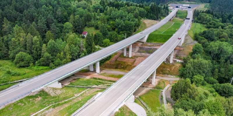 Widok z drona na imponującą estakadę drogi ekspresowej S1 w rejonie Węgierskiej Górki. Konstrukcja przebiega wysoko nad doliną, pośród gęstych lasów i zabudowań. Widoczne ekrany akustyczne i oznaczenia wykonawcy inwestycji – firmy Mirbud.