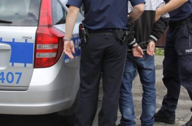 Zatrzymany mężczyzna w kajdankach stojący obok radiowozu policyjnego, pilnowany przez dwóch funkcjonariuszy Policji w umundurowaniu.