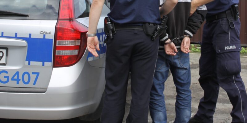Zatrzymany mężczyzna w kajdankach stojący obok radiowozu policyjnego, pilnowany przez dwóch funkcjonariuszy Policji w umundurowaniu.