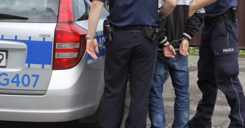 Zatrzymany mężczyzna w kajdankach stojący obok radiowozu policyjnego, pilnowany przez dwóch funkcjonariuszy Policji w umundurowaniu.