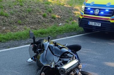 Motocykl leżący na jezdni po kolizji drogowej, obok pojazd ratownictwa medycznego z włączonymi światłami sygnalizacyjnymi. Zdarzenie miało miejsce na tle zalesionej okolicy w Grojcu, w powiecie oświęcimskim.