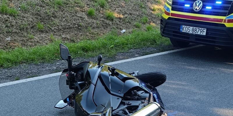 Motocykl leżący na jezdni po kolizji drogowej, obok pojazd ratownictwa medycznego z włączonymi światłami sygnalizacyjnymi. Zdarzenie miało miejsce na tle zalesionej okolicy w Grojcu, w powiecie oświęcimskim.