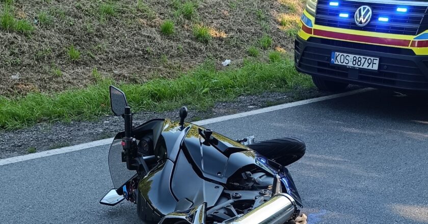 Motocykl leżący na jezdni po kolizji drogowej, obok pojazd ratownictwa medycznego z włączonymi światłami sygnalizacyjnymi. Zdarzenie miało miejsce na tle zalesionej okolicy w Grojcu, w powiecie oświęcimskim.