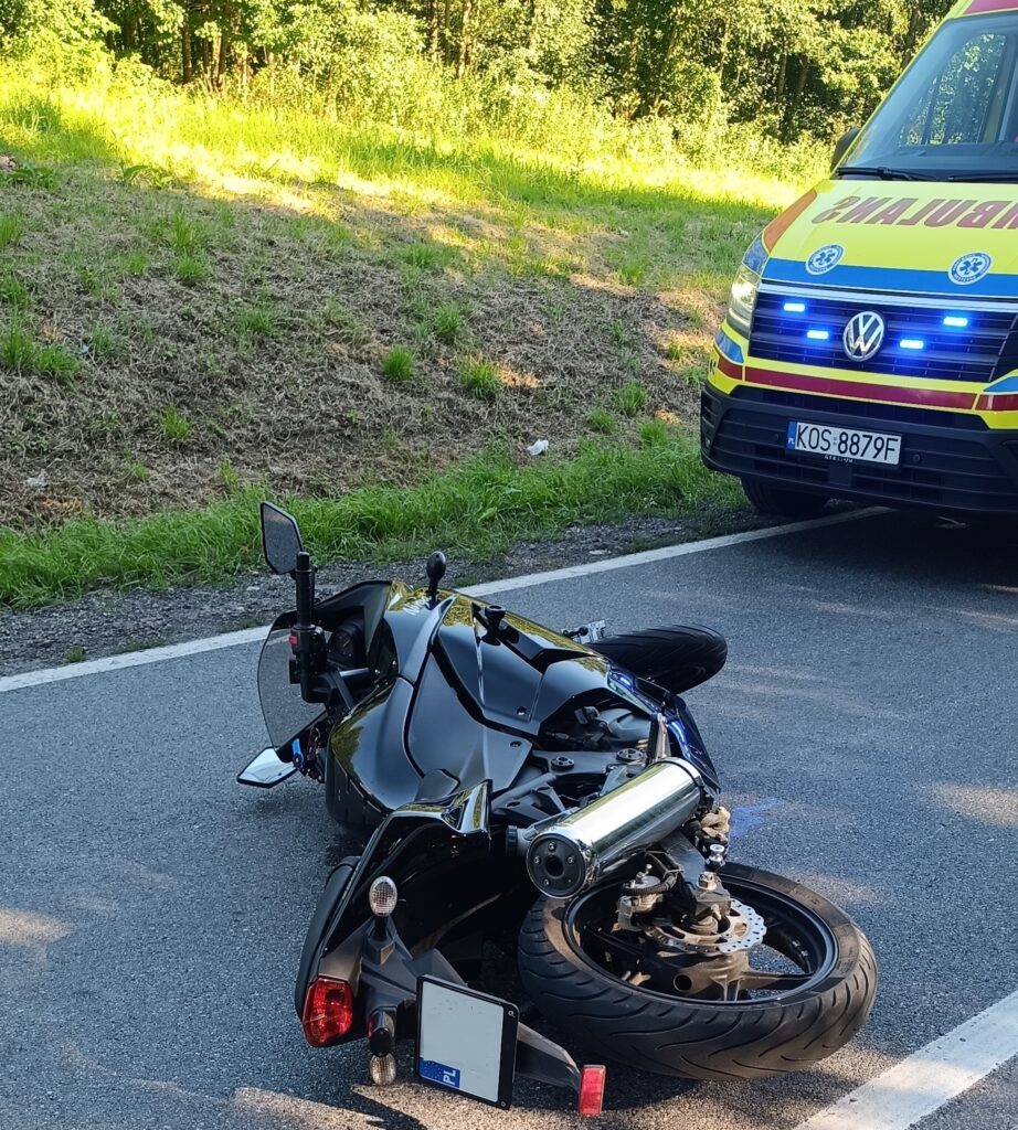 Motocykl leżący na jezdni po kolizji drogowej, obok pojazd ratownictwa medycznego z włączonymi światłami sygnalizacyjnymi. Zdarzenie miało miejsce na tle zalesionej okolicy w Grojcu, w powiecie oświęcimskim.