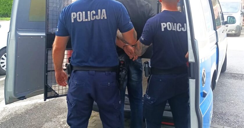 Policjanci z Oświęcimia prowadzą zatrzymanego mężczyznę w kajdankach do radiowozu.