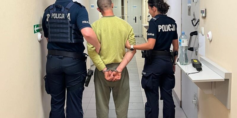 Dwóch policjantów – kobieta i mężczyzna – prowadzi skute kajdankami mężczyznę korytarzem w budynku komendy Policji. Funkcjonariusze idą obok zatrzymanego, który ma ręce skute