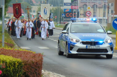 Radiowóz policyjny pilotuje pieszą pielgrzymkę z udziałem duchownych i wiernych, maszerujących ulicami Oświęcimia z kościoła św. Maksymiliana. W tle widoczne szyldy sklepów i centrum handlowego.