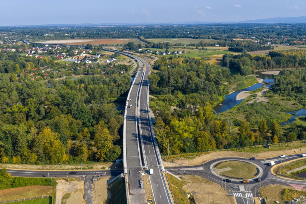 Widok z drona na nowo wybudowany odcinek południowej obwodnicy Oświęcimia – most nad rzeką Sołą i rondo turbinowe, otoczone zielenią i zabudową jednorodzinną. Trwa budowa drugiej nitki jezdni.