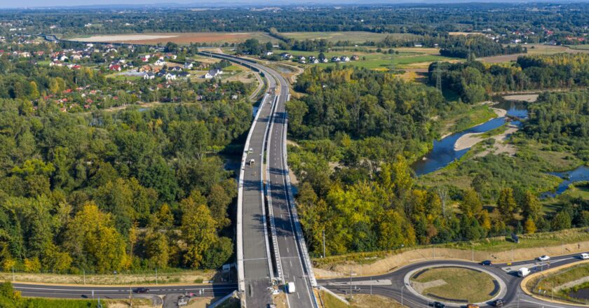 Widok z drona na nowo wybudowany odcinek południowej obwodnicy Oświęcimia – most nad rzeką Sołą i rondo turbinowe, otoczone zielenią i zabudową jednorodzinną. Trwa budowa drugiej nitki jezdni.