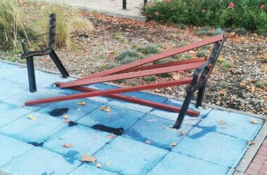 Zniszczona ławka na placu zabaw w Oświęcimiu – złamane deski leżą na podłożu, metalowa konstrukcja wygięta. W tle rower, zieleń i grupa mieszkańców siedzących na innej ławce.