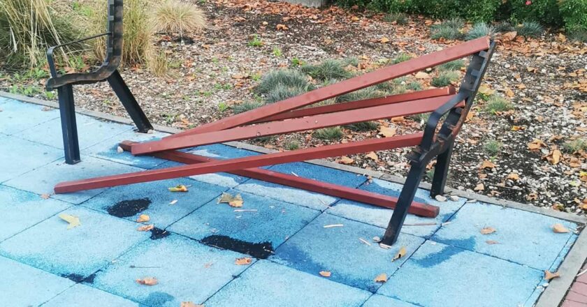 Zniszczona ławka na placu zabaw w Oświęcimiu – złamane deski leżą na podłożu, metalowa konstrukcja wygięta. W tle rower, zieleń i grupa mieszkańców siedzących na innej ławce.