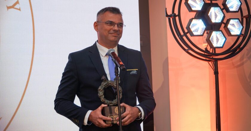 Na zdjęciu widać Radosław Włoszek prezes Koleje Małopolskie, mężczyznę w garniturze i niebieskim krawacie, stojącego przy mikrofonie. W rękach trzyma statuetkę nagrody Grossi Cracovienses. Za nim znajduje się fragment scenografii gali – jasne tło oraz nowoczesna lampa sceniczna emitująca niebieskie światło.