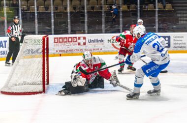 Zawodnik Re-Plast Unii Oświęcim (numer 21, Takulja) oddaje strzał na bramkę podczas meczu hokeja na lodzie przeciwko drużynie w czerwono-zielonych strojach. Bramkarz rywali wykonuje paradę obronną, próbując zatrzymać krążek. Sędzia obserwuje akcję z boku lodowiska.