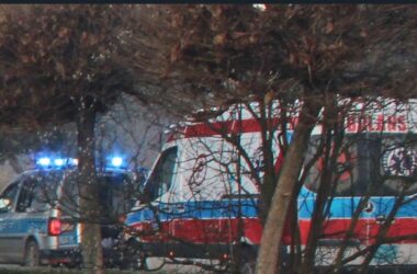 Radiowóz policyjny na sygnałach świetlnych obok karetki pogotowia podczas wspólnej interwencji, widok zza drzew