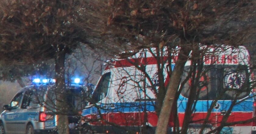 Radiowóz policyjny na sygnałach świetlnych obok karetki pogotowia podczas wspólnej interwencji, widok zza drzew