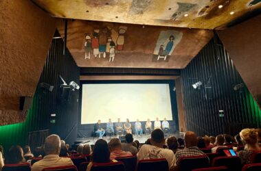 Publiczność wypełnia salę Oświęcimskiego Centrum Kultury podczas premiery filmu „Rok 2020” Piotra Struczyka; na scenie przed ekranem siedzą twórcy i aktorzy, a sufit zdobi charakterystyczne malowidło z postaciami w styluPubliczność wypełnia salę Oświęcimskiego Centrum Kultury podczas premiery filmu „Rok 2020” Piotra Struczyka; na scenie przed ekranem siedzą twórcy i aktorzy, a sufit zdobi charakterystyczne malowidło z postaciami w stylu nawiązującym do sztuki współczesnej. nawiązującym do sztuki współczesnej.