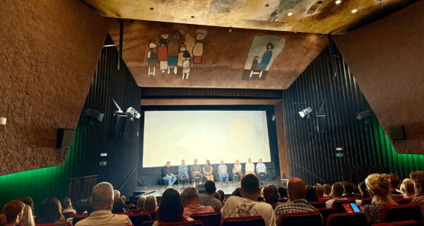 Publiczność wypełnia salę Oświęcimskiego Centrum Kultury podczas premiery filmu „Rok 2020” Piotra Struczyka; na scenie przed ekranem siedzą twórcy i aktorzy, a sufit zdobi charakterystyczne malowidło z postaciami w styluPubliczność wypełnia salę Oświęcimskiego Centrum Kultury podczas premiery filmu „Rok 2020” Piotra Struczyka; na scenie przed ekranem siedzą twórcy i aktorzy, a sufit zdobi charakterystyczne malowidło z postaciami w stylu nawiązującym do sztuki współczesnej. nawiązującym do sztuki współczesnej.